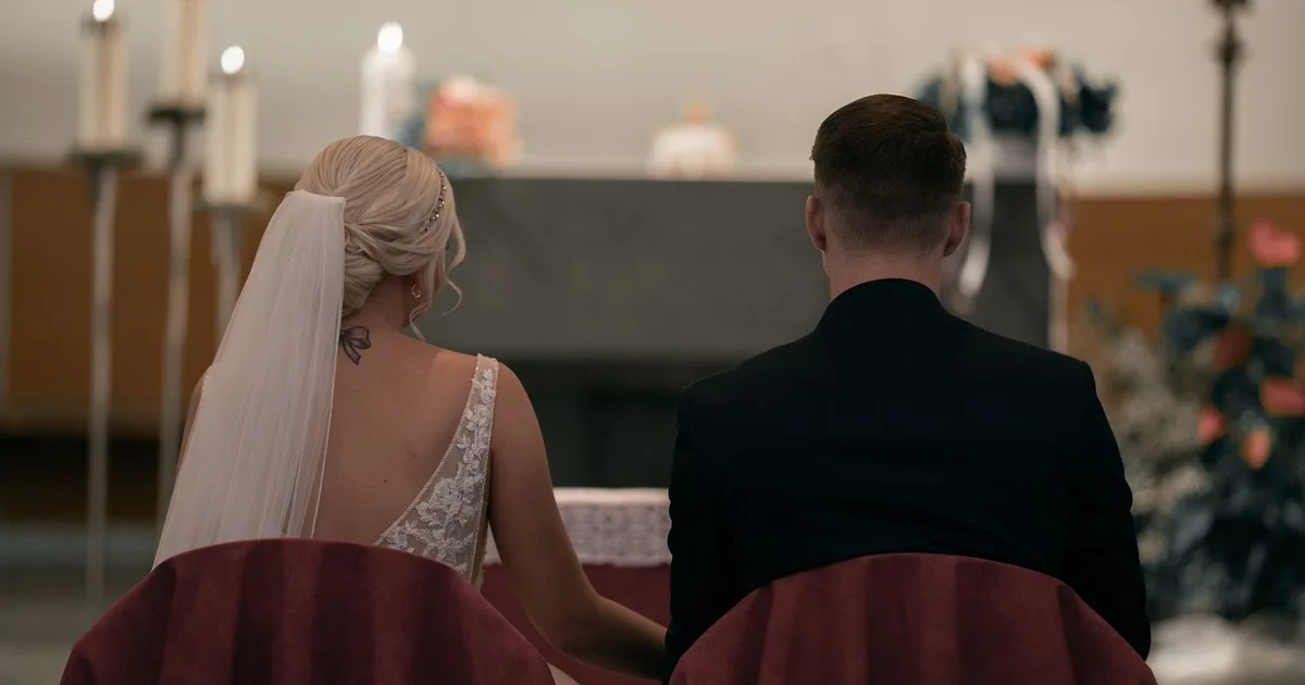 Brautpaar von hinten, auf Stühlen vor dem Altar einer Kirche zwischen Kerzenleuchtern sitzend