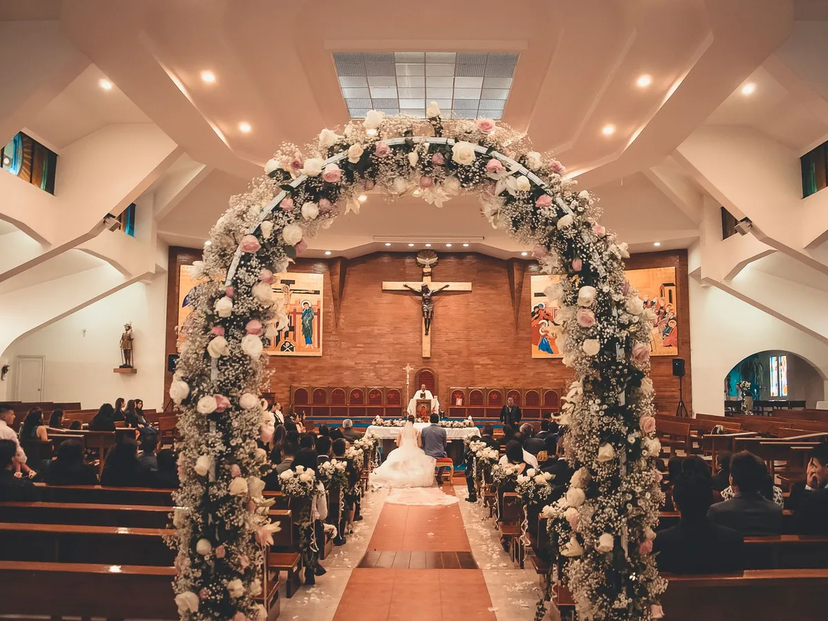 Blick durch einen Blumenbogen ins Kirchenschiff, Gäste in den Bänken, das Brautpaar sitzt vorn vor dem Altar