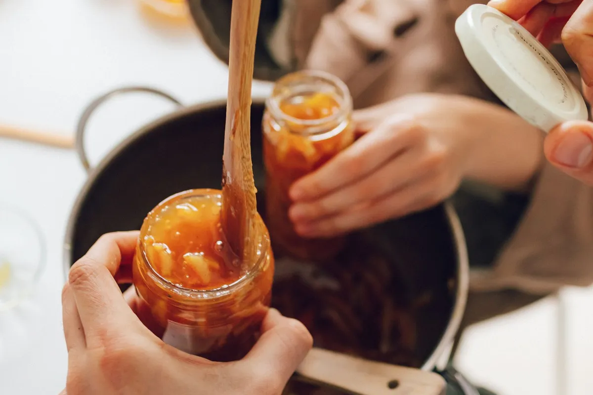 Zwei Personen füllen mit einem Holzlöffel Orangenmarmelade aus einem Topf in Schraubgläser, eine Hand hält den Deckel bereit