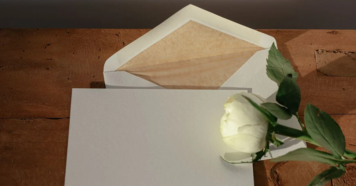 Weißes Kärtchen mit Briefumschlag und weißer Rose auf einem Holztisch, bereit für einen Glückwunsch