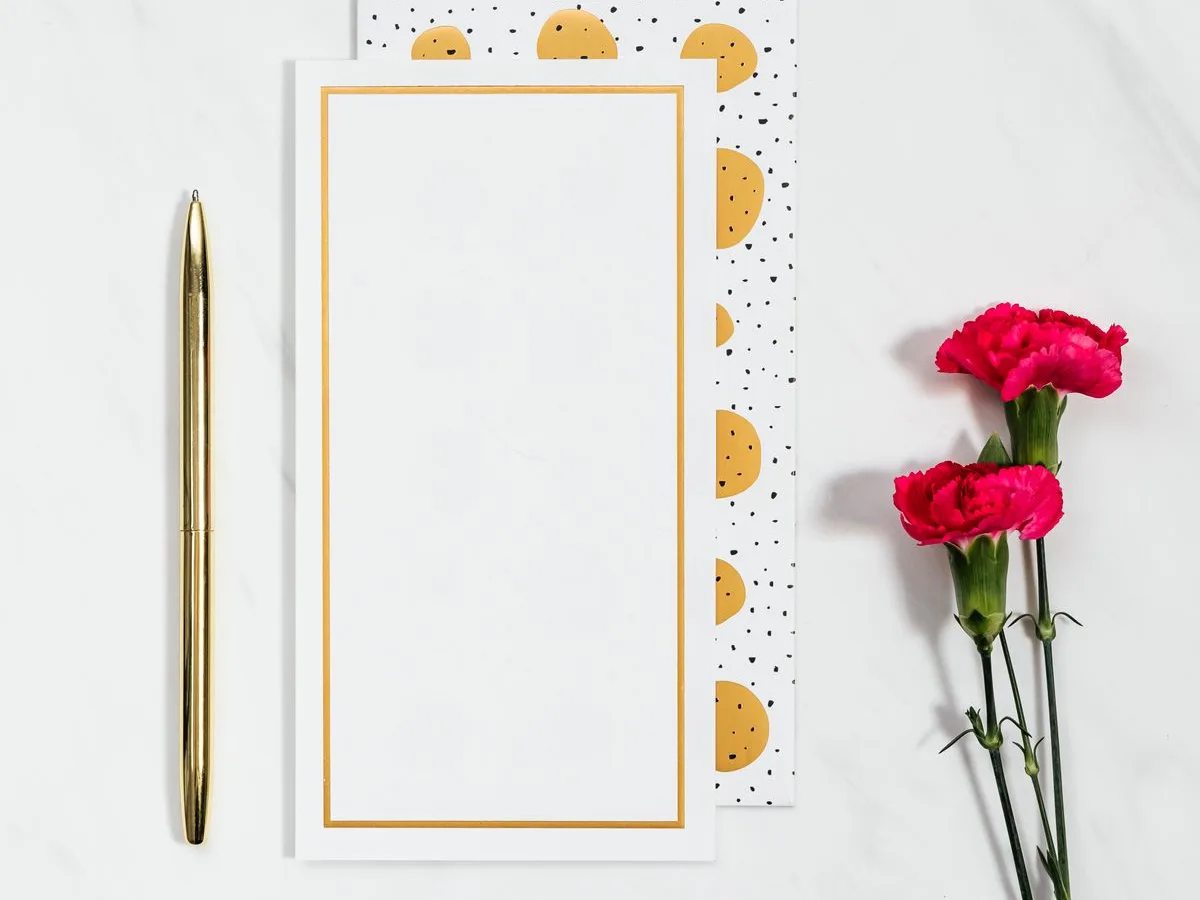Goldener Stift neben einer leeren Glückwunschkarte mit goldenem Rahmen und pinken Nelken