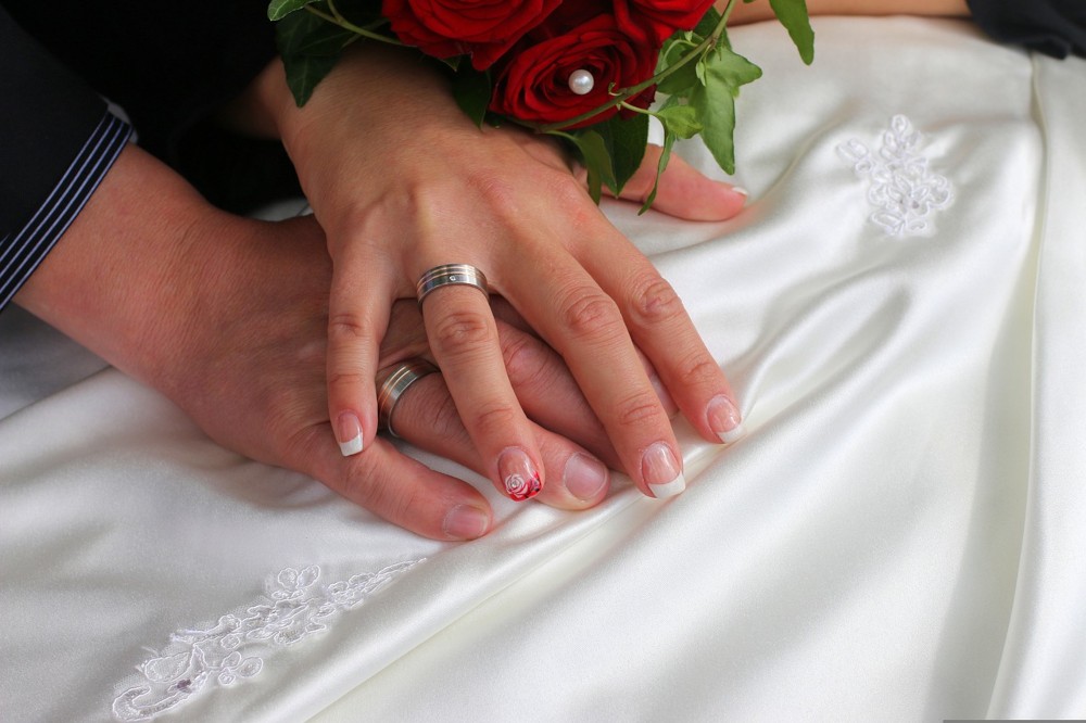 Die Hände von der Braut und dem Bräutigam liegen aufeinander auf dem Brautkleid, während beide Trauringe tragen