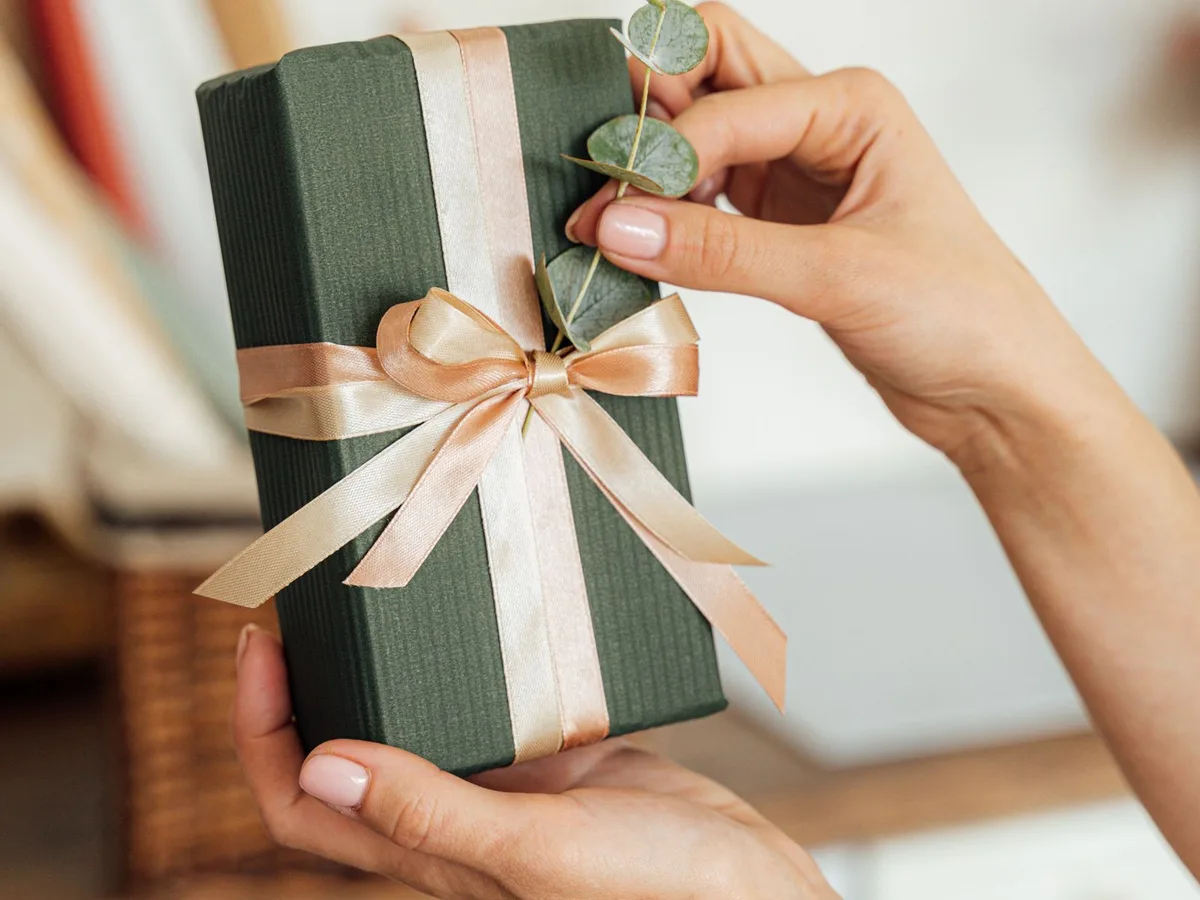 Zwei Hände halten eine dunkelgrün verpackte Geschenkbox mit Satinschleife und Eukalyptuszweig