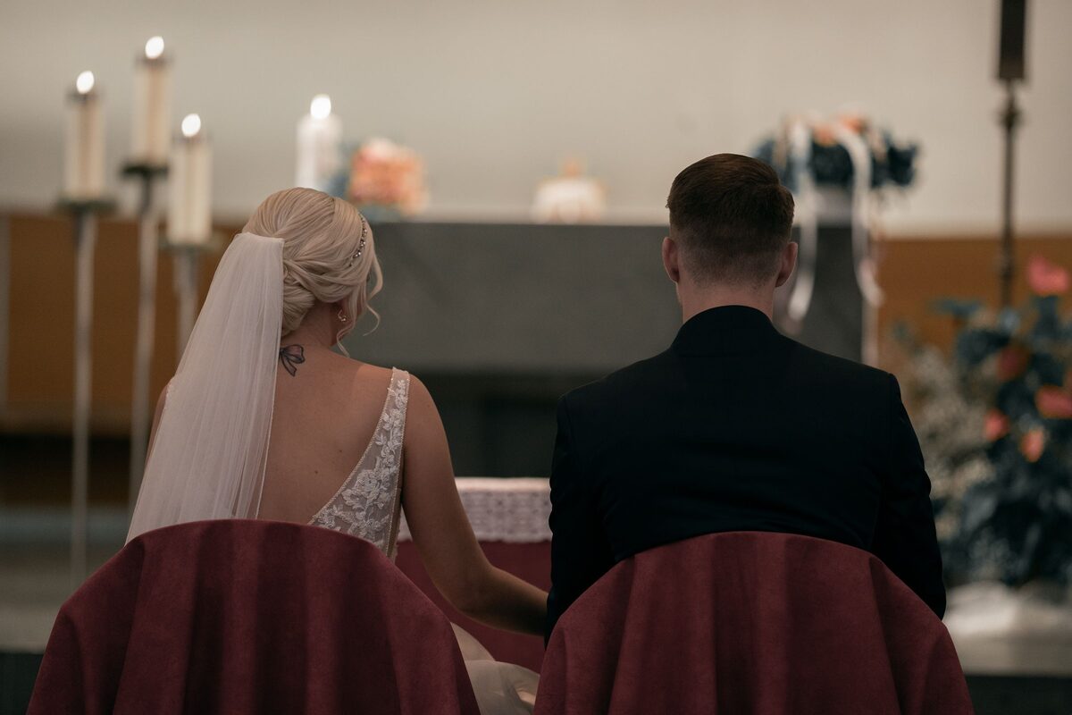 Brautpaar von hinten, auf Stühlen vor dem Altar einer Kirche zwischen Kerzenleuchtern sitzend