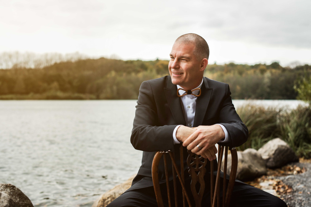 Jens Köhne sitzt im Anzug auf einem Stuhl vor einem See