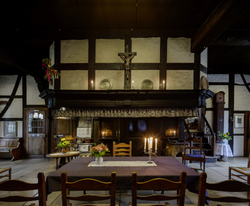 Vorne steht ein großer Tisch für die Trauung, im Hintergrund befindet sich der Kamin und oben ist das Fachwerkgebilde mit einem Kreuz; rechts vom Kamin befindet sich ein Aufgang und eine Standuhr und links ist eine Tür und eine Bank