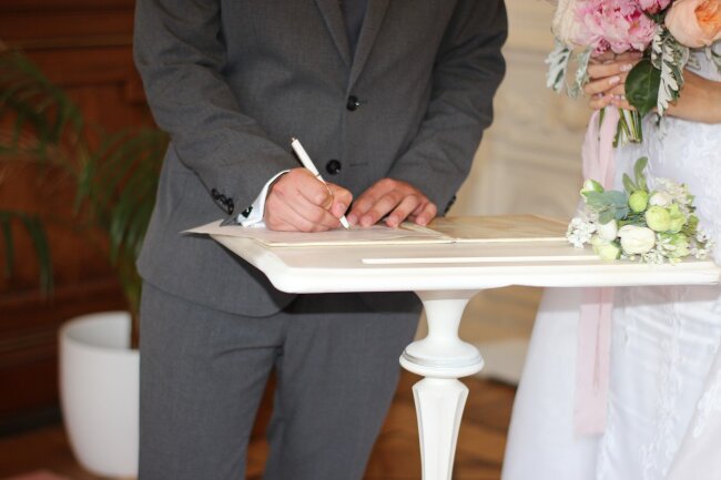 Der Bräutigam unterschreibt die Heiratsurkunde beim Standesamt, während die Braut mit dem Brautstrauß daneben steht